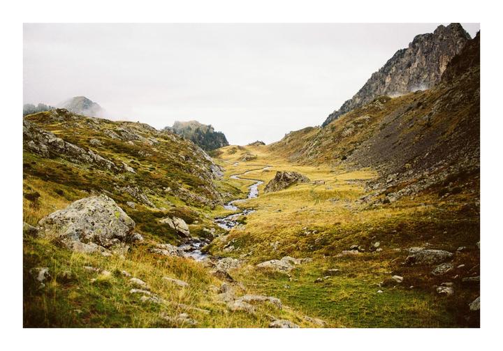 A winding stream flows through a valley surrounded by mountains and rocky, grassy slopes under a hazy sky.