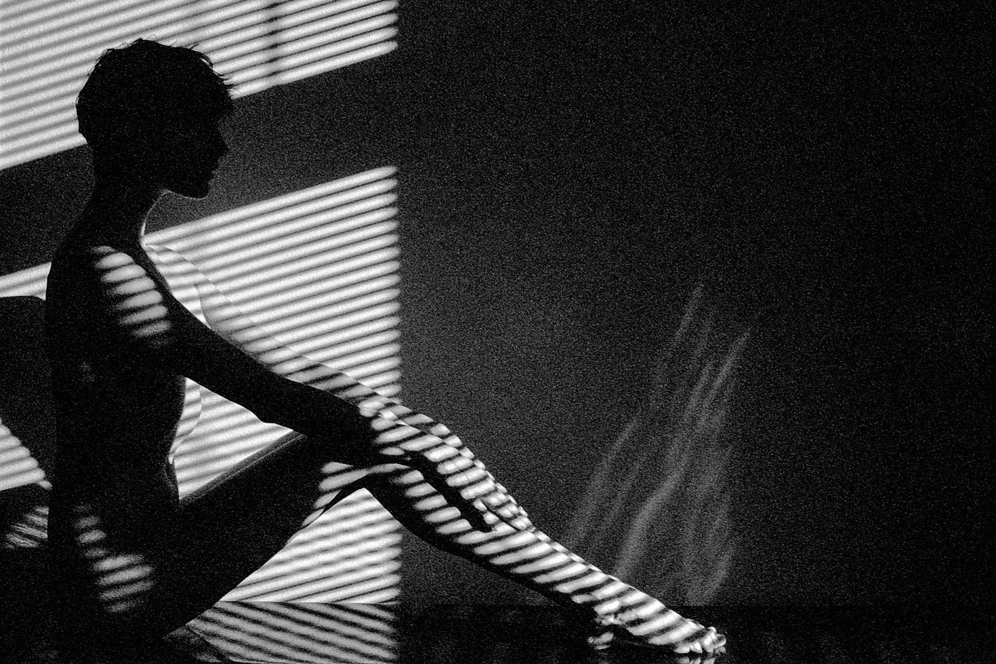 A woman sits in profile with shadows from blinds cast across her body and the wall behind her.