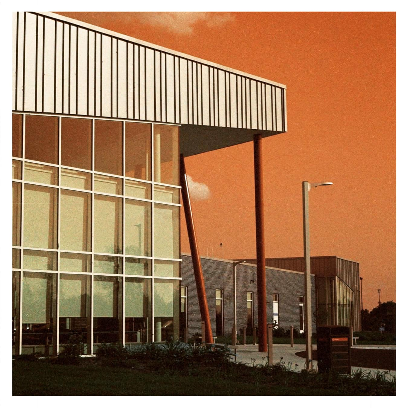 A modern building with large windows and a slatted overhang stands against an orange sky with a single cloud.