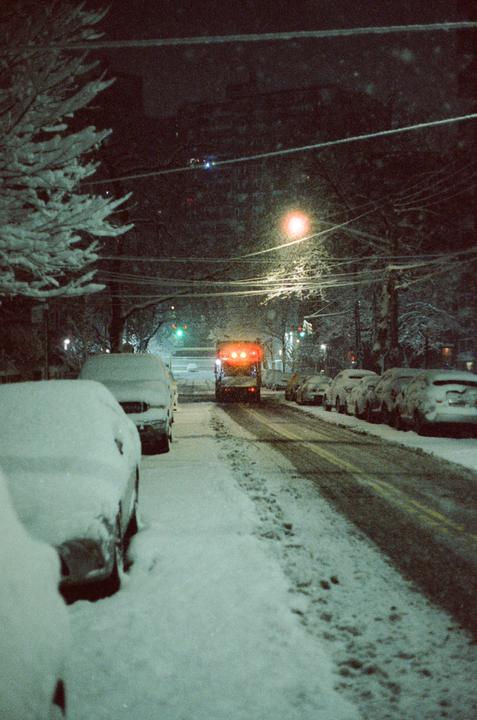 A snow-covered street at night is lined with parked cars and a vehicle with flashing red lights.