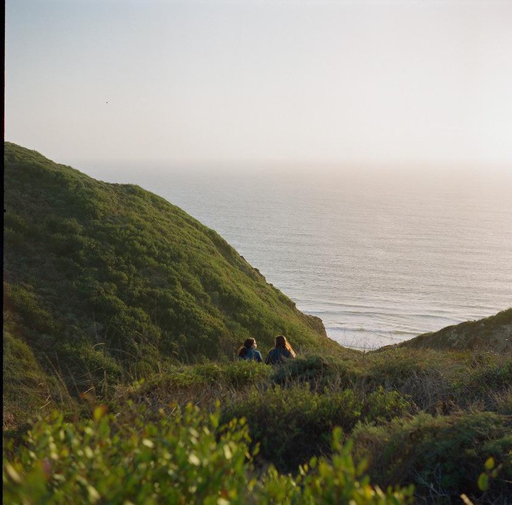 Two people sit on a grassy hillside overlooking the ocean at sunset.