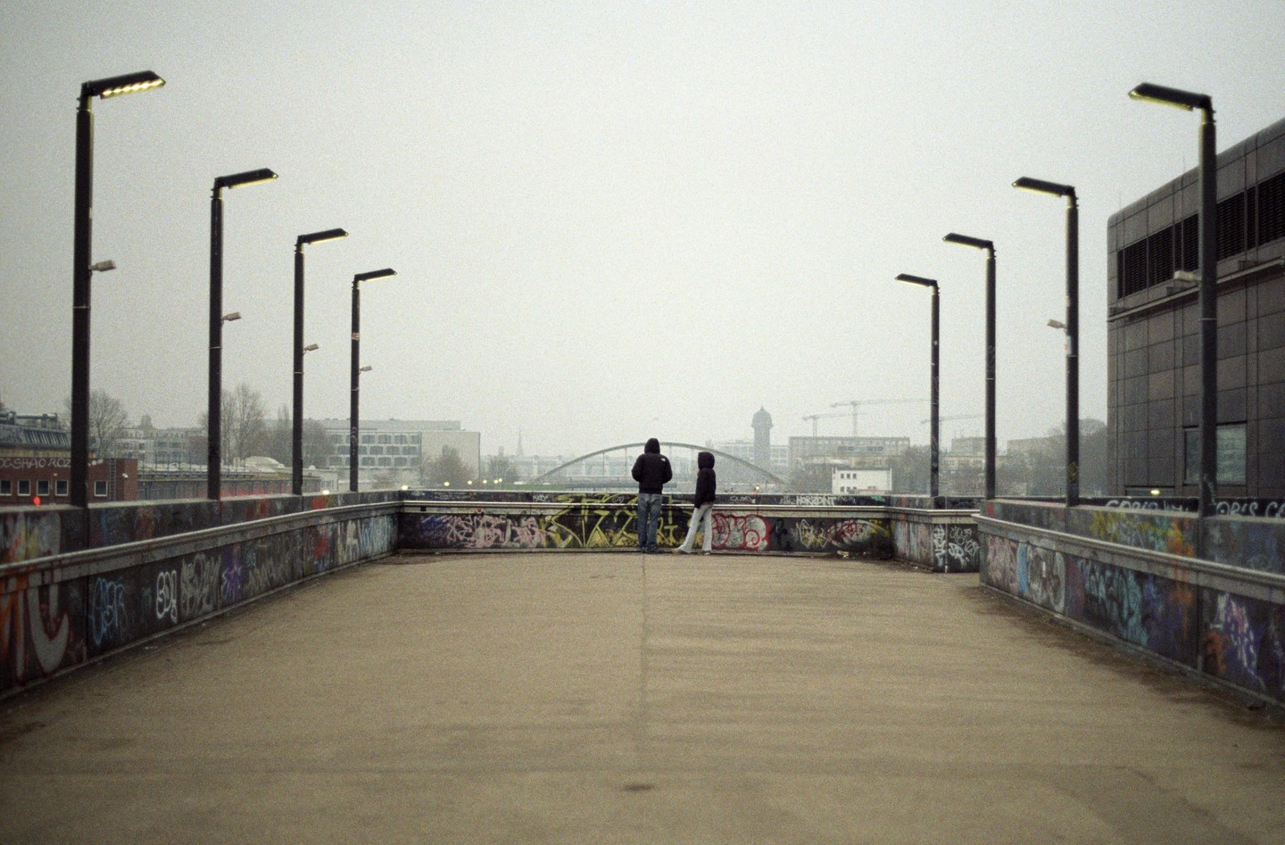 Two people stand on a graffiti-covered bridge, looking out at a city skyline under a hazy sky.