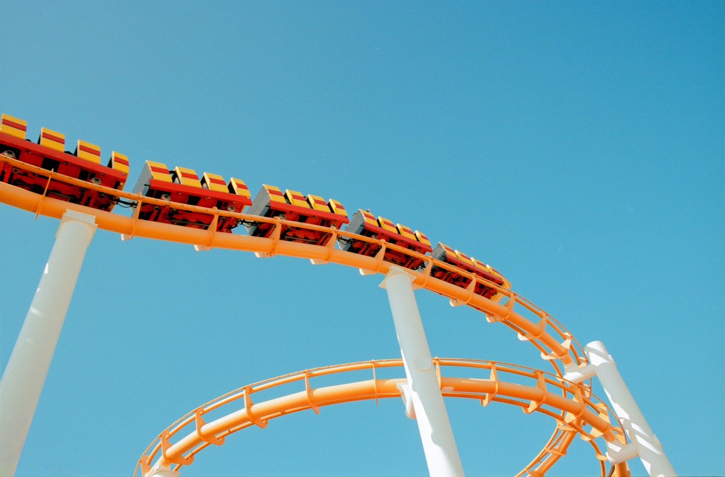 A bright orange roller coaster track with red and yellow cars curves against a clear blue sky.