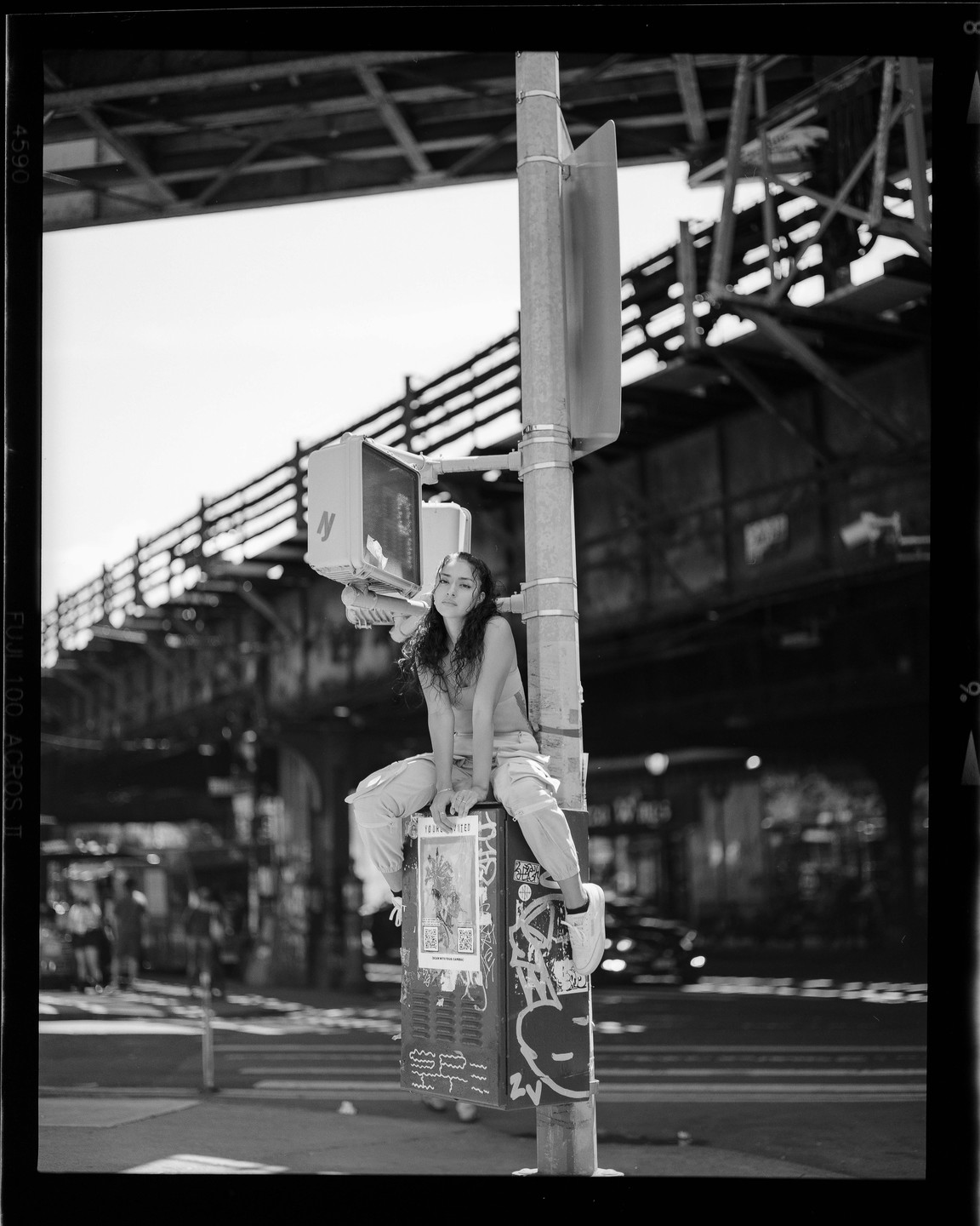 A woman sits on a traffic signal control box on a city street with an elevated train track in the background.