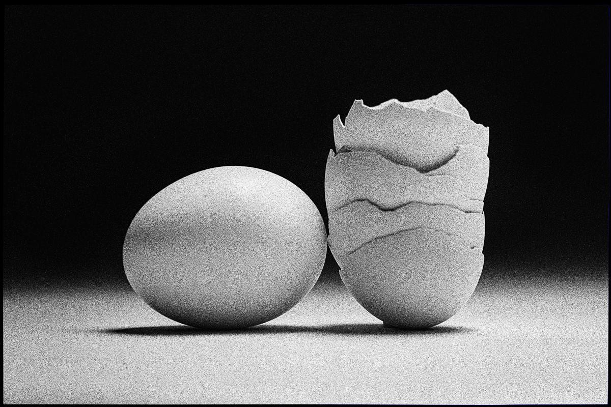 A whole egg sits next to a broken eggshell on a surface with a dark background.