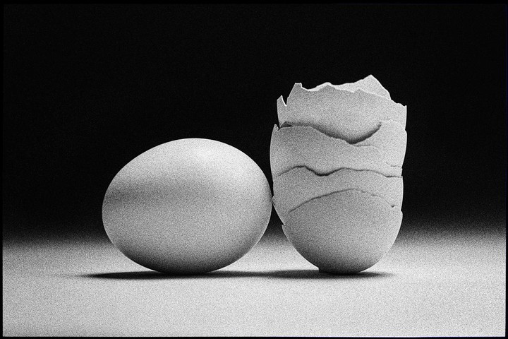 A whole egg sits next to a broken eggshell on a surface with a dark background.