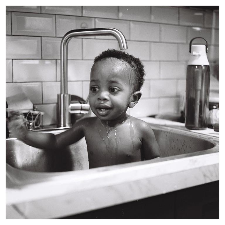 A child is standing in a kitchen sink with water dripping from their hair and chest.