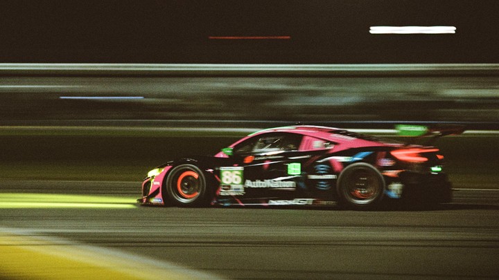 A pink and black race car with the number 86 on its side speeds along a track at night, with motion blur visible.