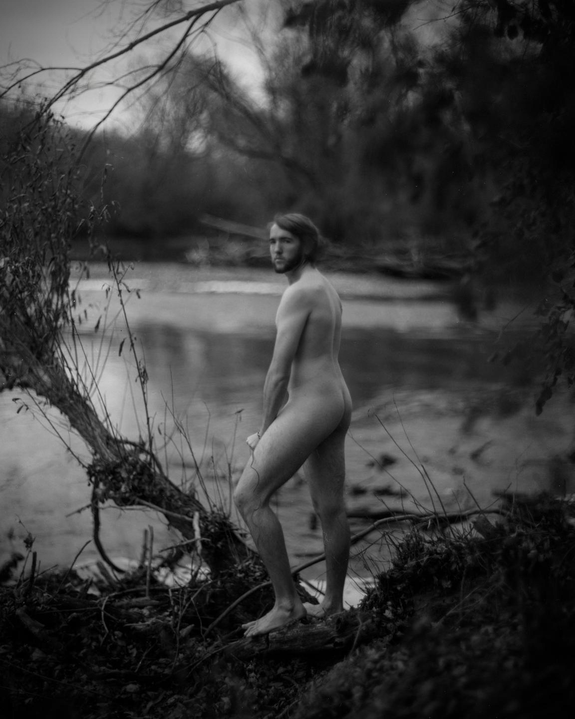 A man stands nude on a riverbank, looking over his shoulder.