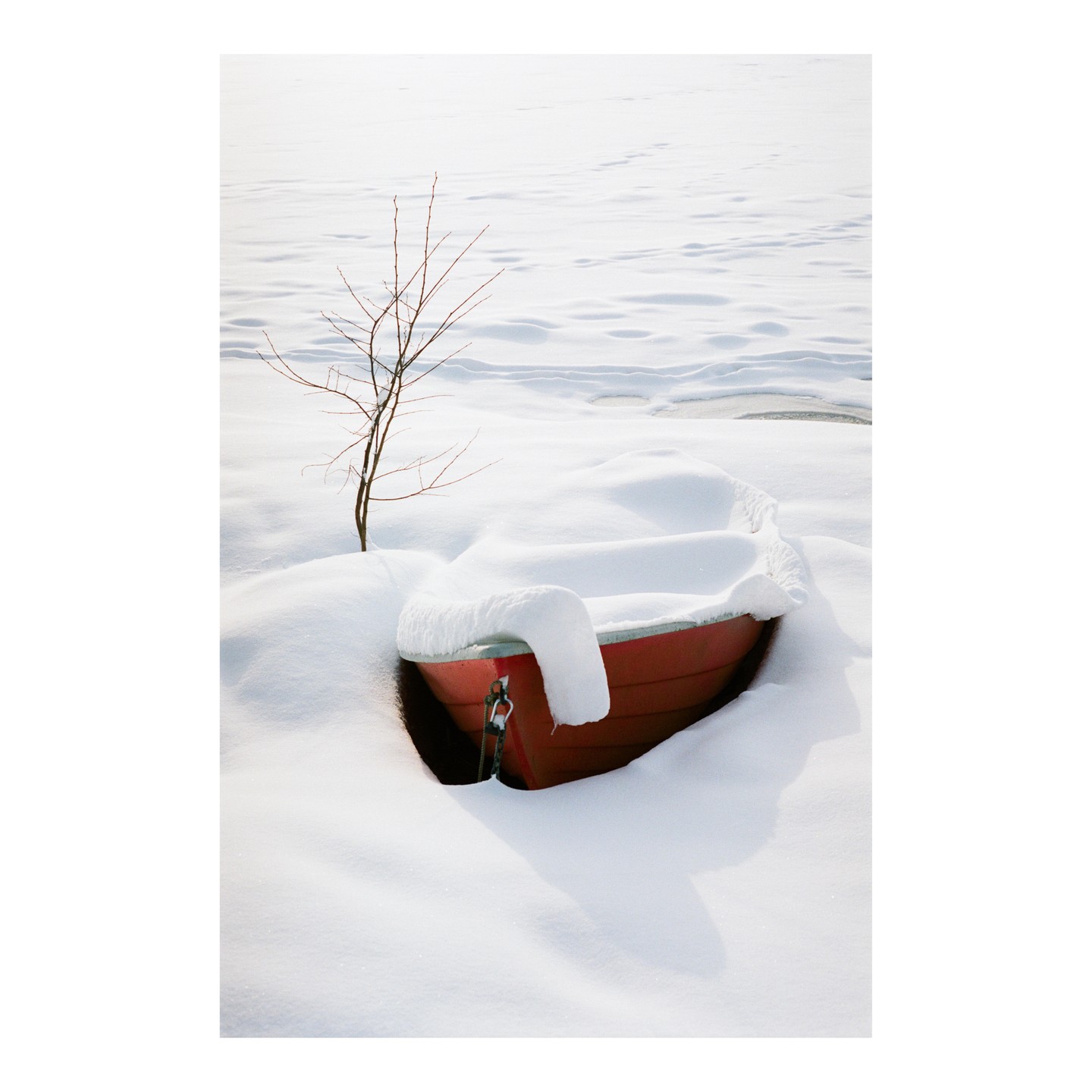 A small red boat is half-buried in snow next to a bare tree on a frozen lake.