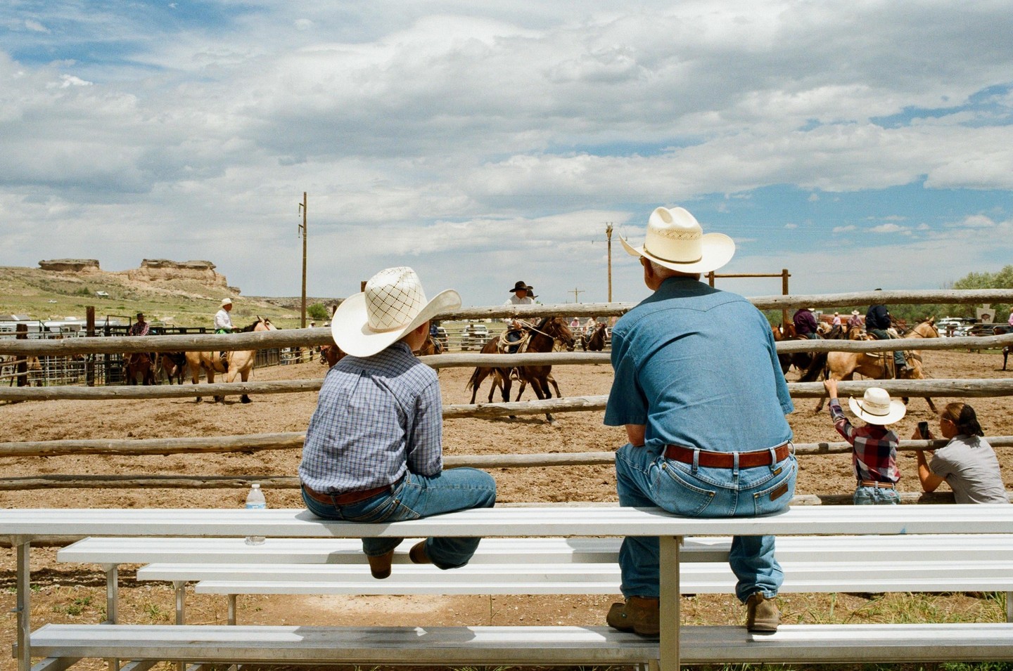 A man and a boy in cowboy hats sit on bleachers watching a rodeo in a dirt arena with horses and riders.