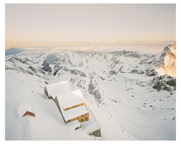 Two buildings with snow-covered roofs sit on a snowy mountain overlooking a valley with more snow-covered mountains and a cloudy sky.