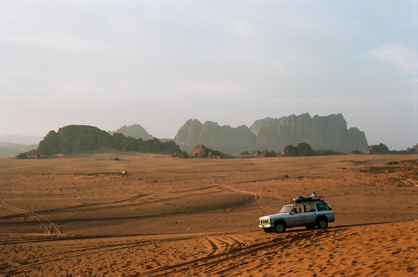 A white SUV with a roof rack drives across a vast desert landscape with mountains in the background.