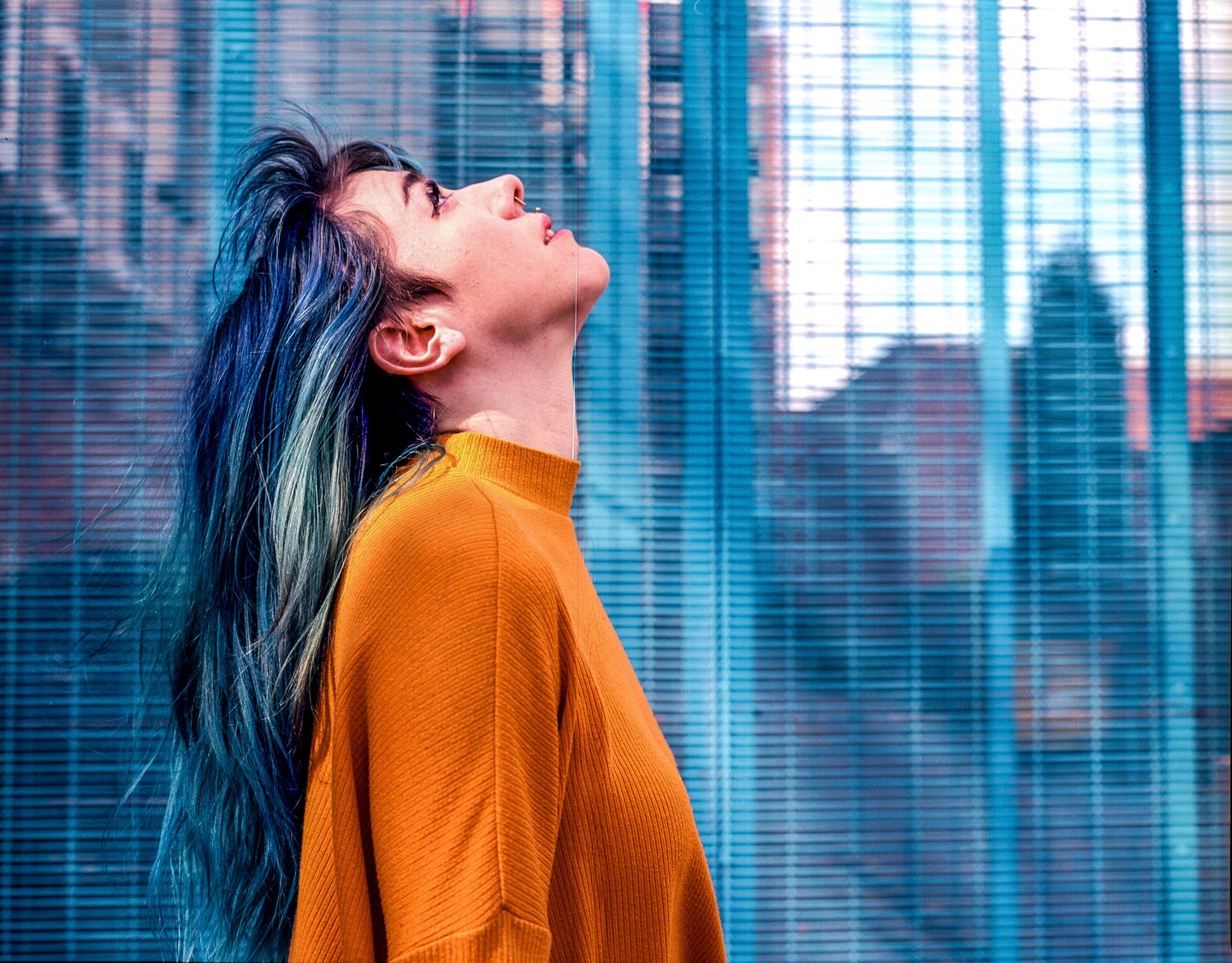 A woman with blue hair and an orange sweater looks up against a blue patterned background.