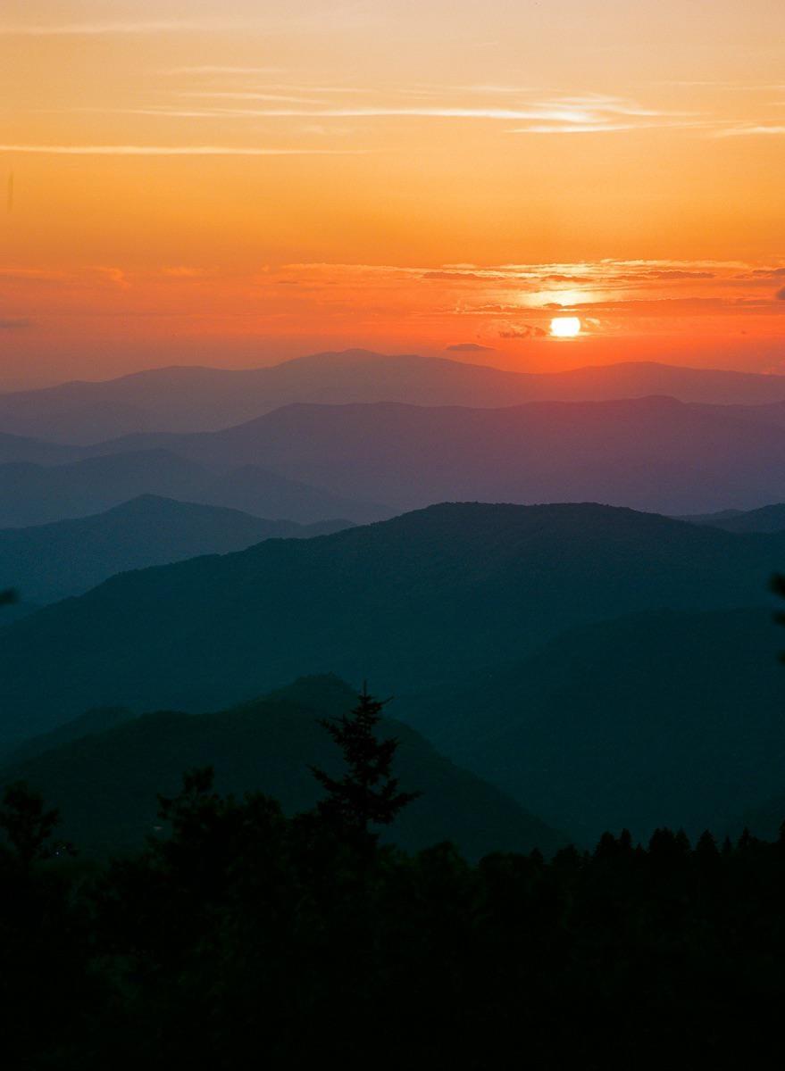 The sun sets over a mountain range with a forest in the foreground.