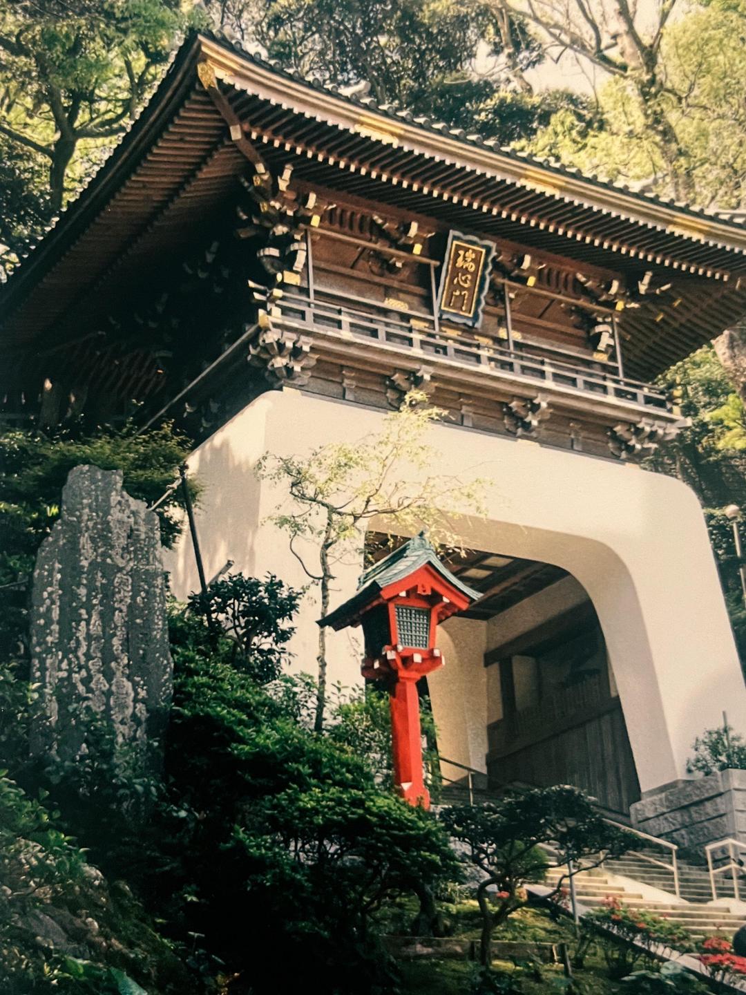 A traditional Japanese temple gate with a red lantern and lush greenery is nestled on a hillside.