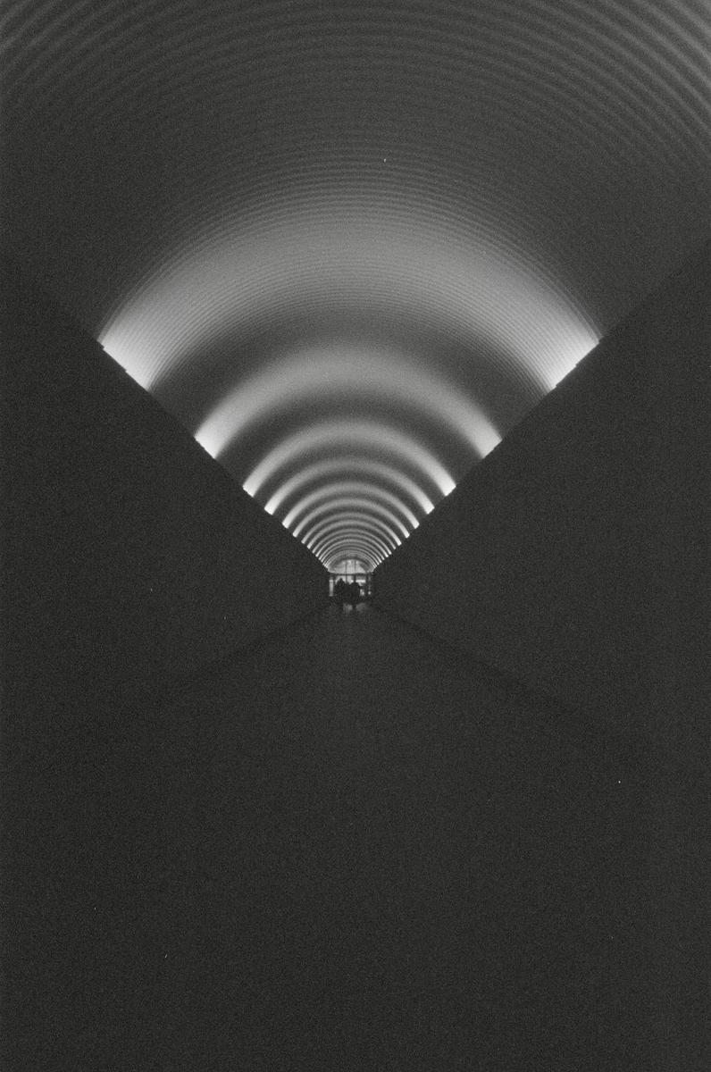 A long, dark, corrugated tunnel is illuminated by a series of lights along its curved ceiling, leading to a distant, small figure.