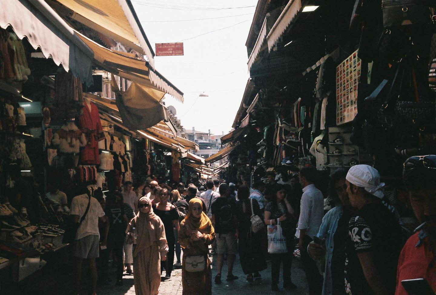 A crowded market street is lined with stalls selling various goods under awnings.