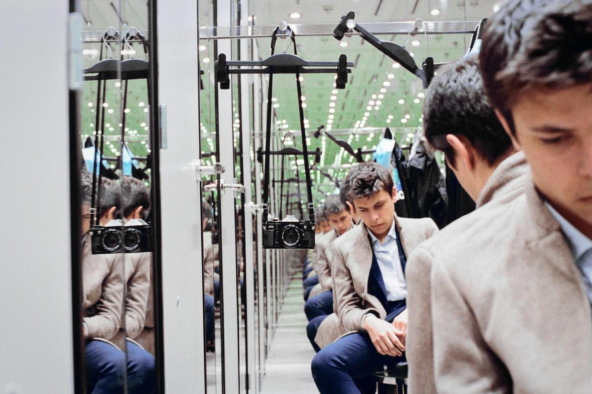 A man sits in a fitting room with mirrors reflecting his image and a camera hanging in the foreground.