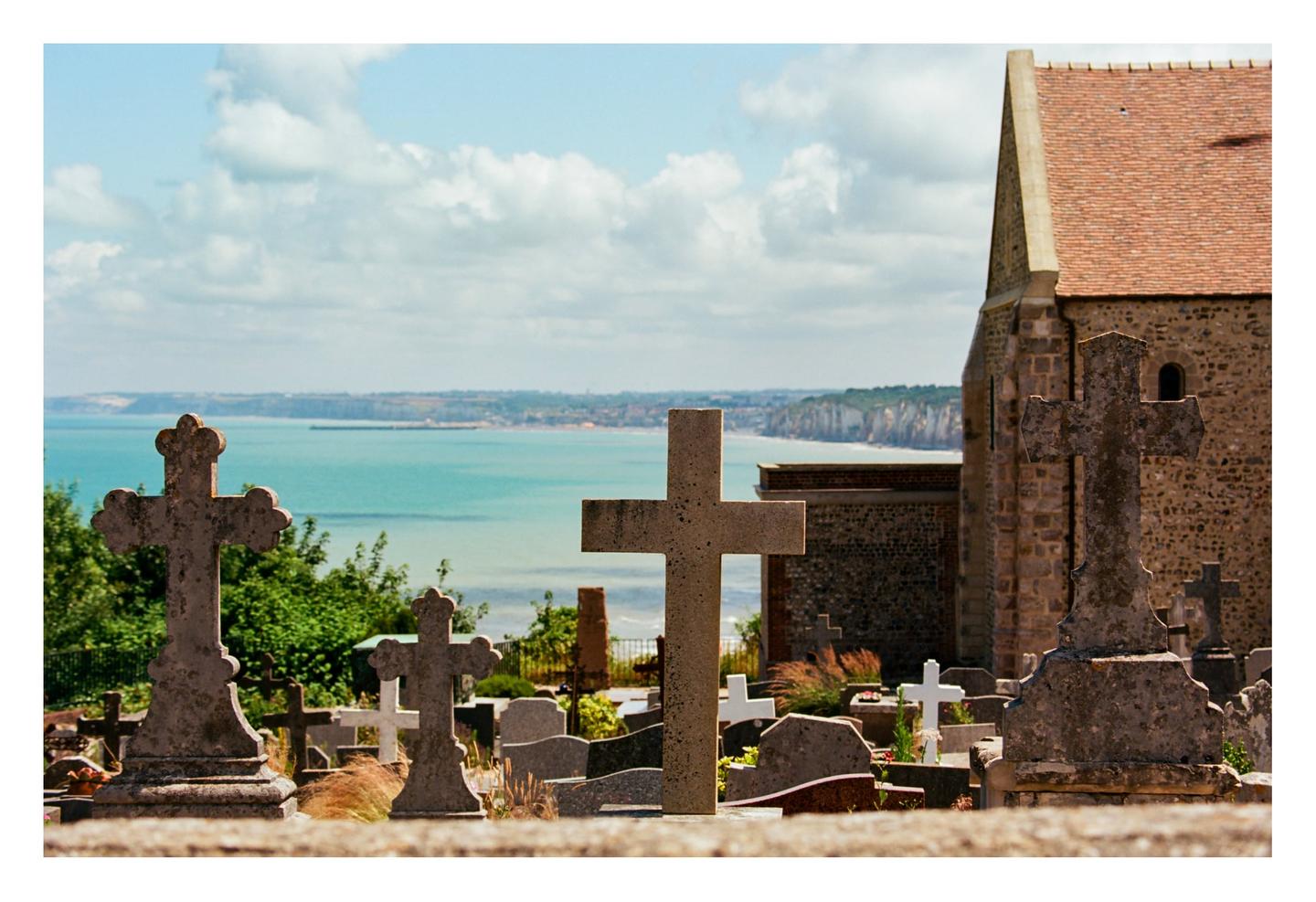 A graveyard with many crosses and tombstones overlooks a body of water and cliffs under a cloudy sky.