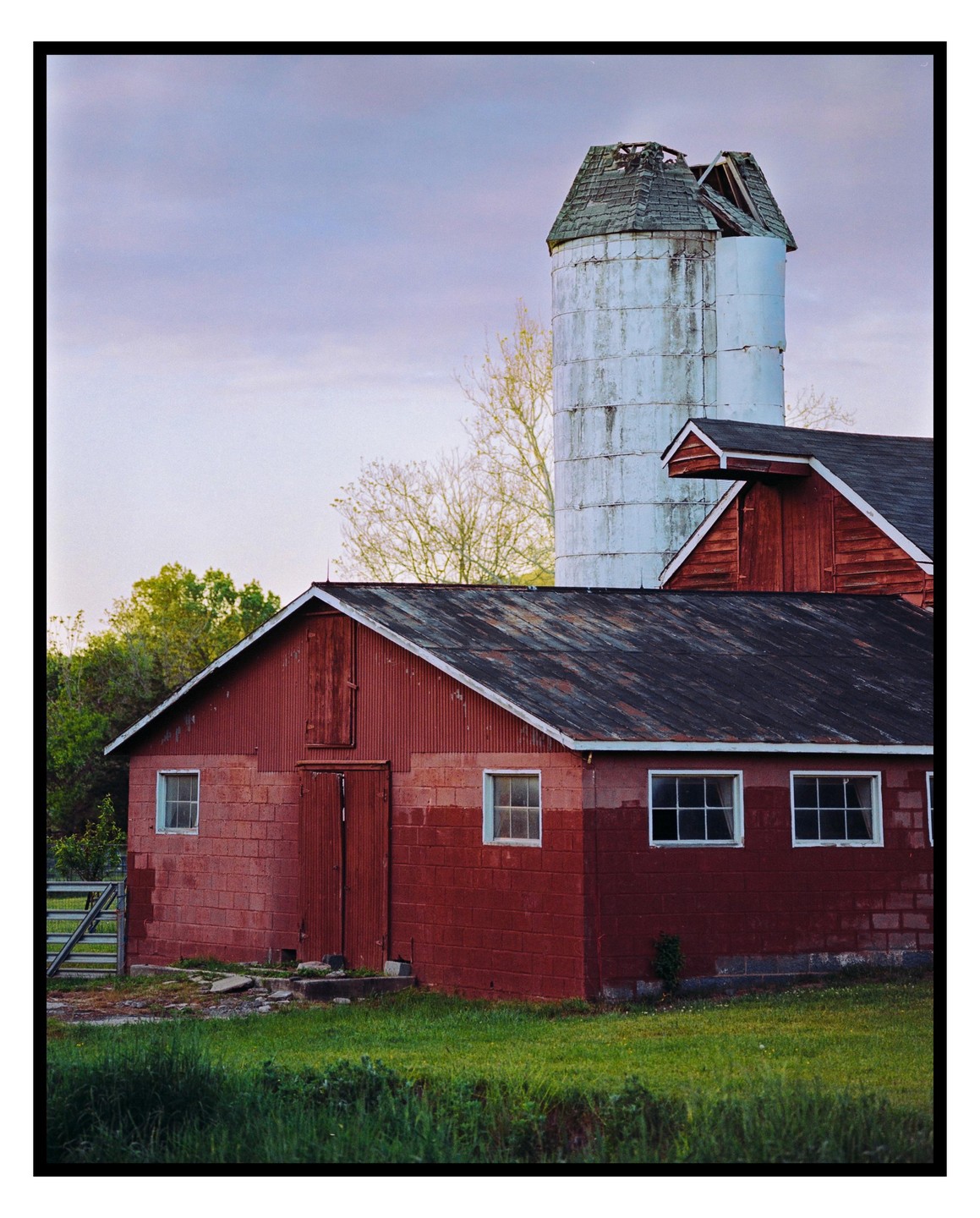A red barn with a white silo stands in a grassy field under a cloudy sky.