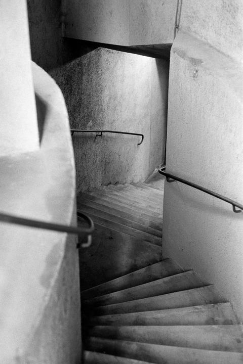 A black and white image shows a concrete spiral staircase with metal handrails descending into shadow.