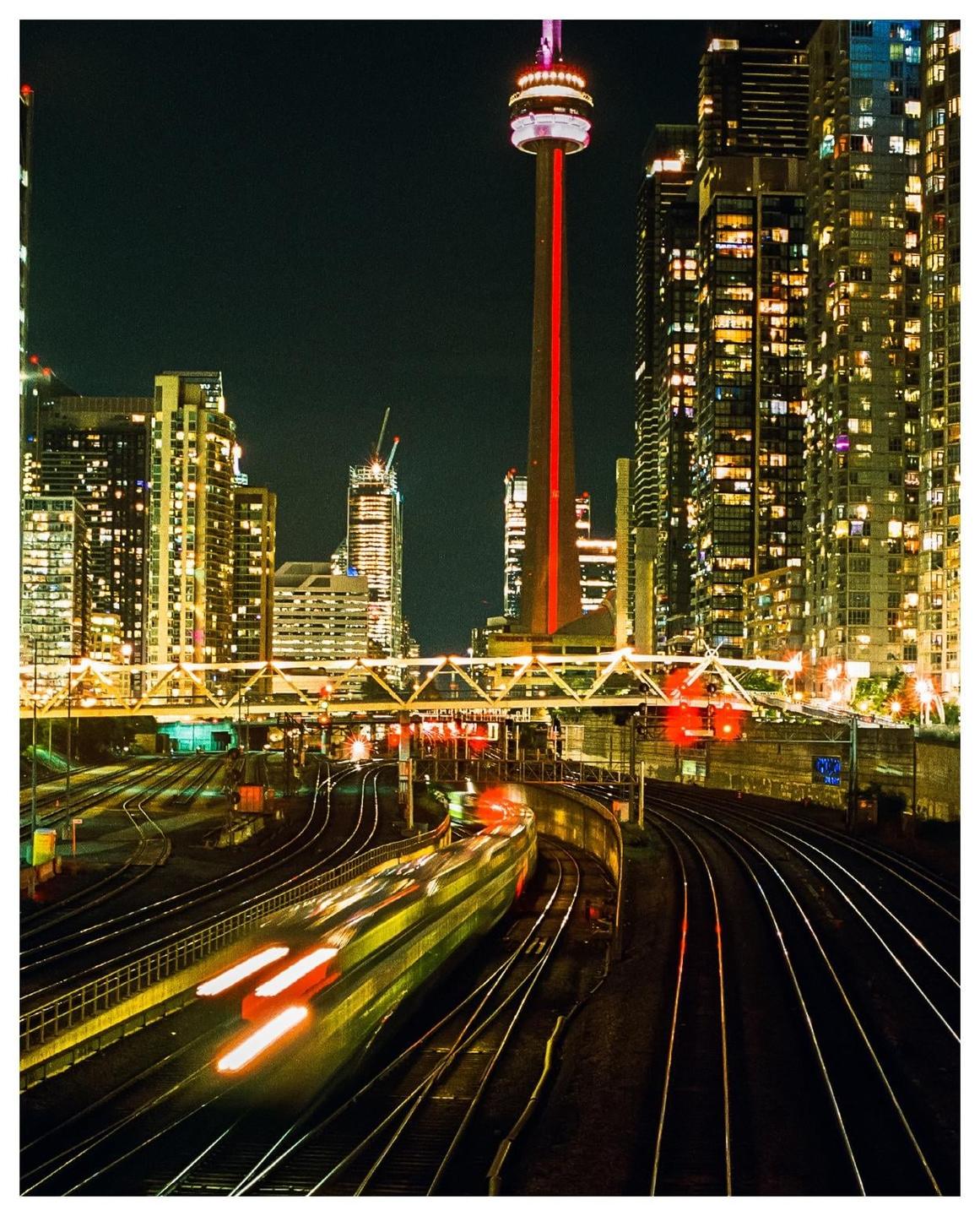A train with light trails moves along tracks at night with a city skyline and the CN Tower in the background.