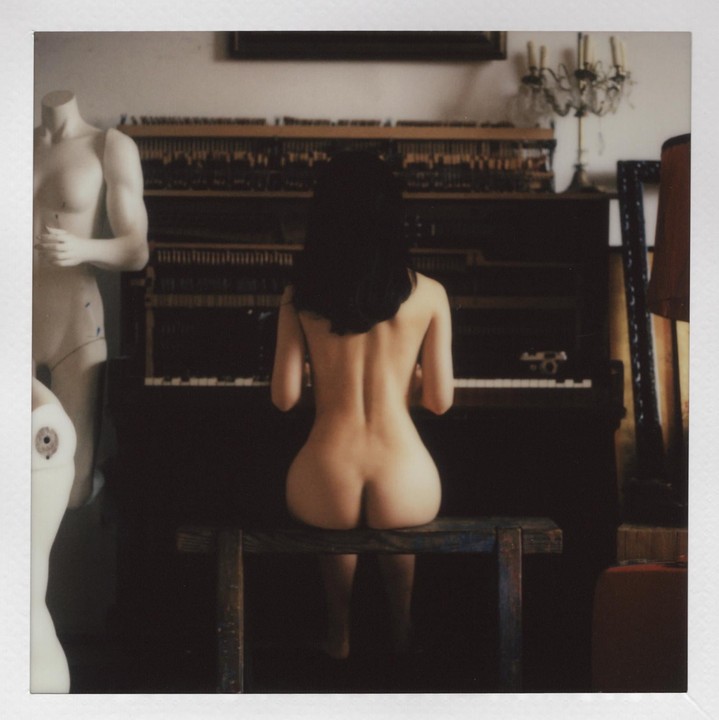 A nude woman sits on a bench with her back to the viewer, playing a piano.