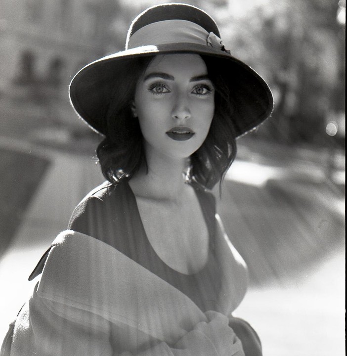 A woman wearing a wide-brimmed hat and a coat looks directly at the viewer.
