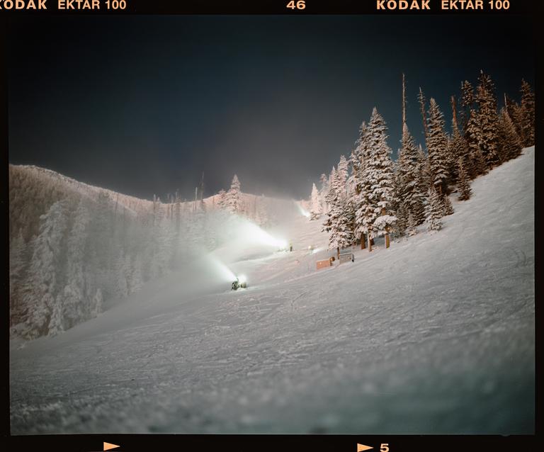 Snow-covered pine trees line a ski slope where snowmaking machines are operating at night.