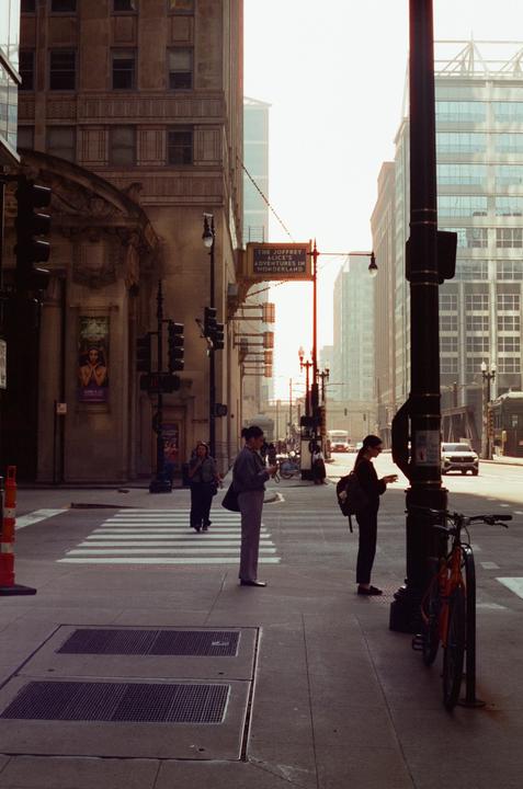 Three women are standing on a city street corner with tall buildings and a street sign in the background.