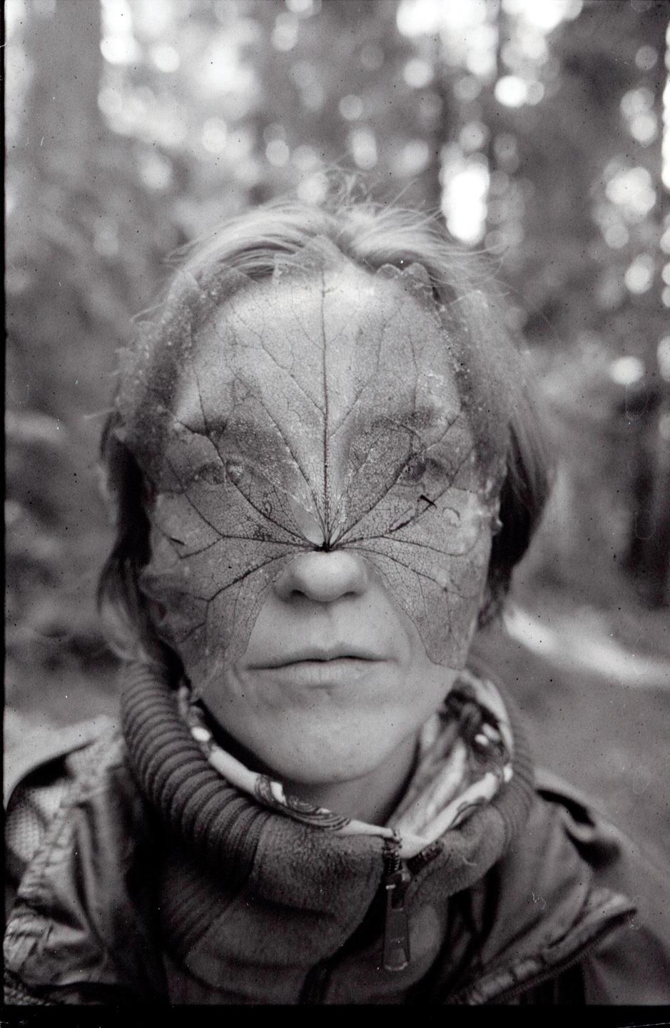 A woman with a large leaf covering her eyes and nose looks directly at the viewer.