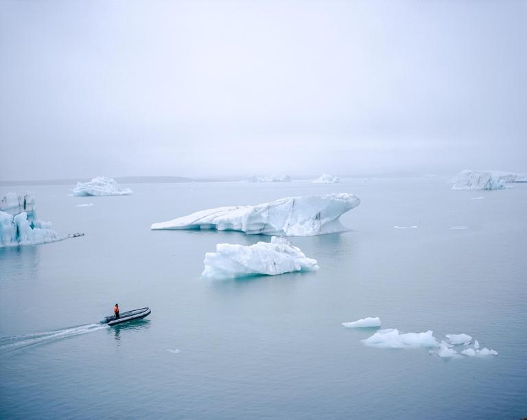 A small boat with a person in an orange jacket navigates through a body of water filled with icebergs under a hazy sky.