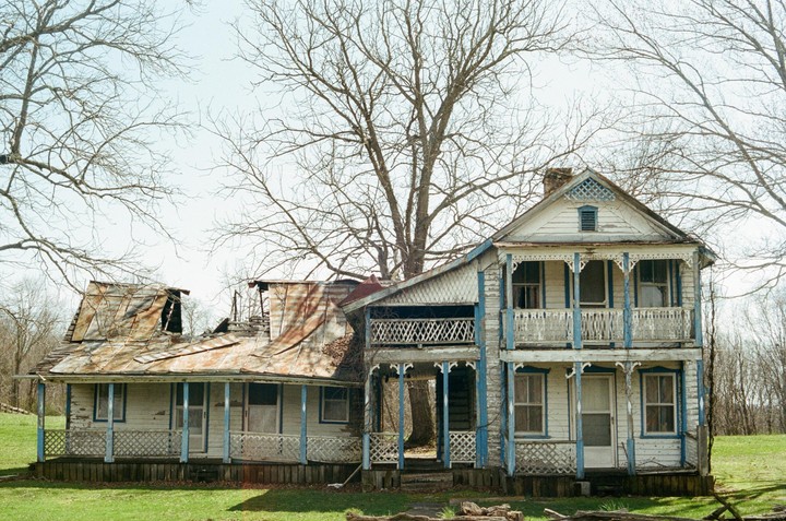 Two dilapidated houses with peeling paint and damaged roofs stand in a grassy field under a clear sky, surrounded by bare trees.