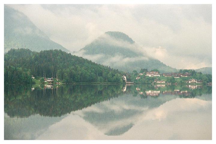 A village with houses and trees is reflected in a lake with mountains in the background.