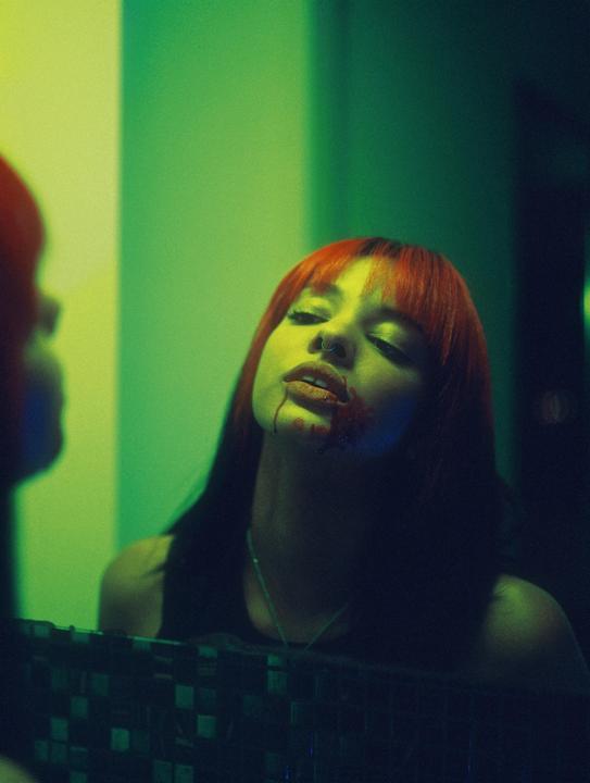 A woman with red hair and blood on her mouth looks up at her reflection in a mirror.