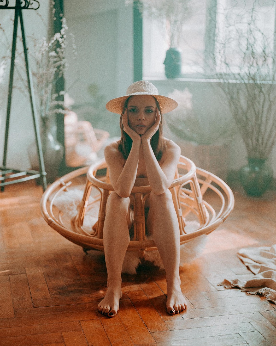 A woman wearing a straw hat sits in a round wicker chair with her hands on her chin.