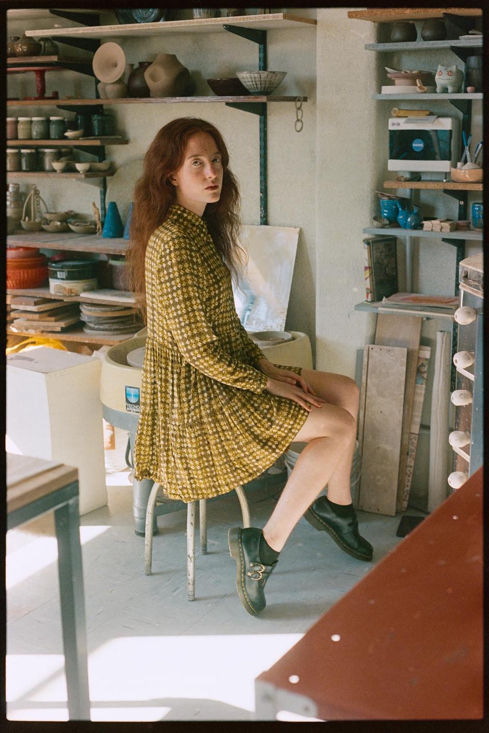 A woman with long red hair sits on a stool in a pottery studio, looking over her shoulder at the viewer.