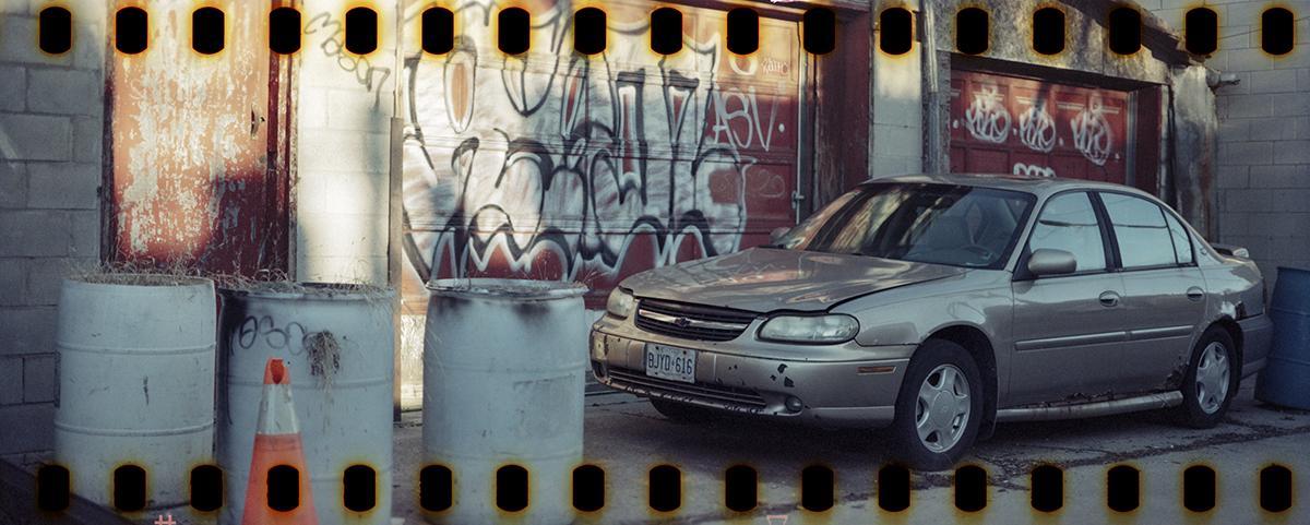 A silver sedan is parked in a back alley next to several barrels and a building with graffiti-covered doors.