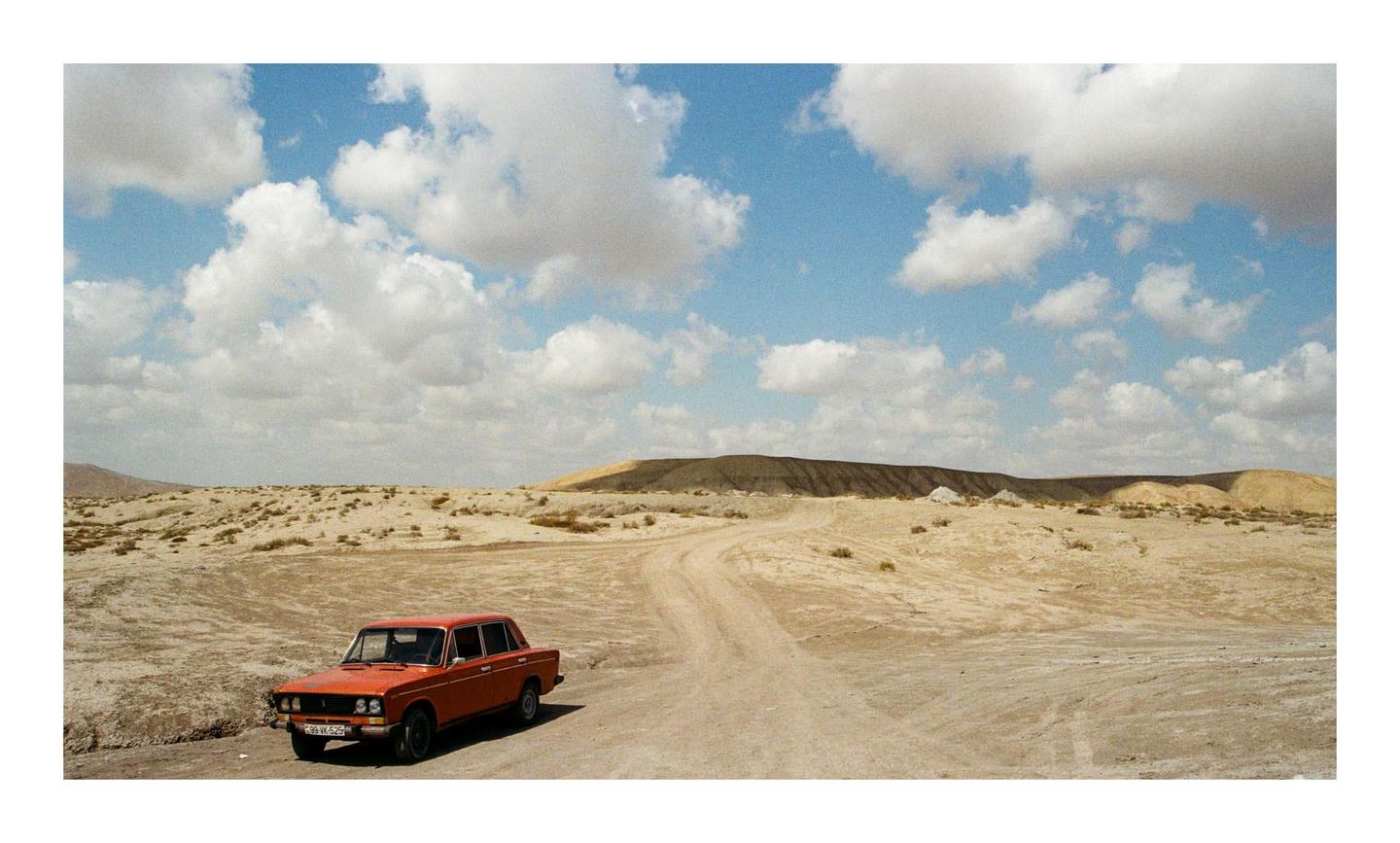 An orange car is parked on a dirt road in a desert landscape under a blue sky with clouds.