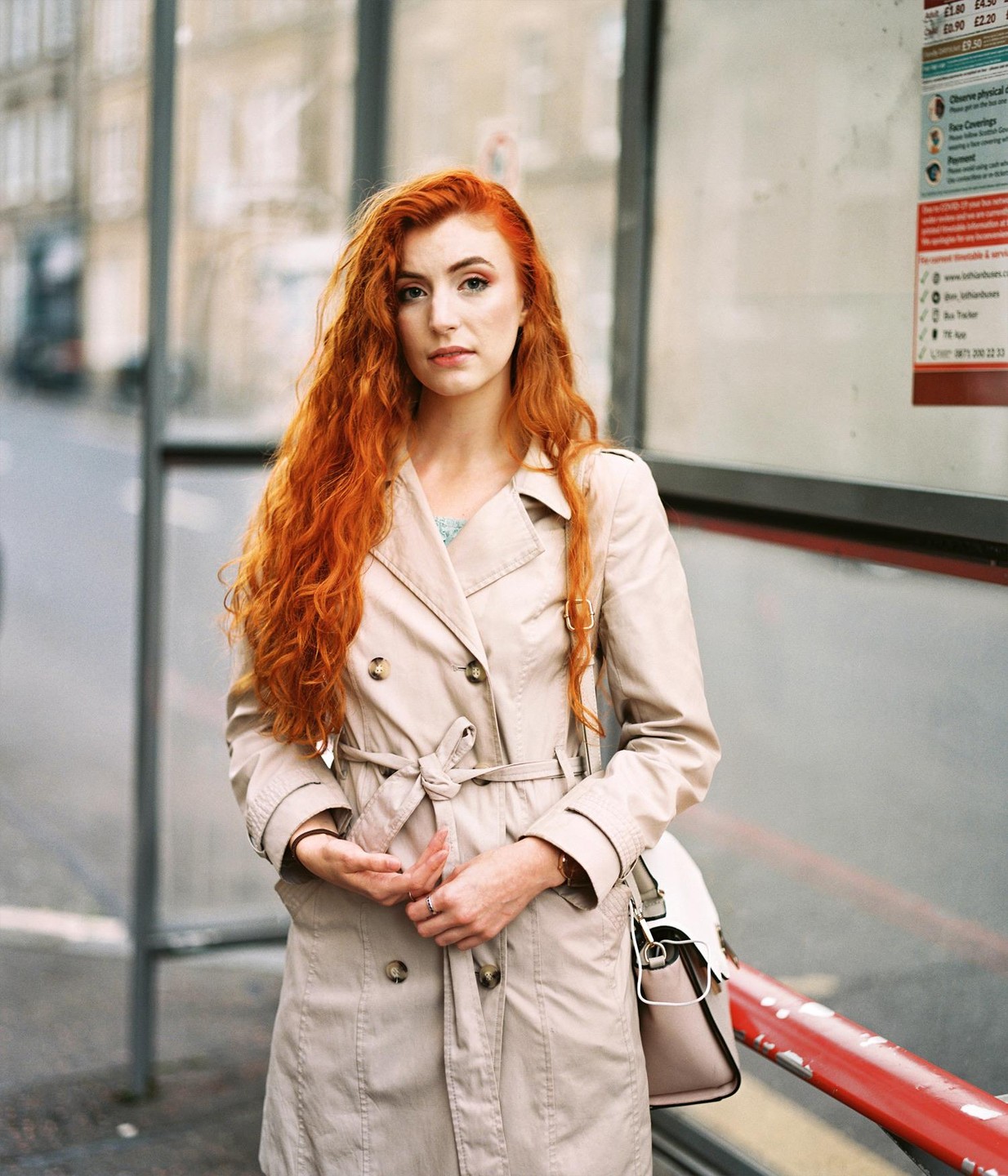 A woman with long red hair and a trench coat stands at a bus stop.