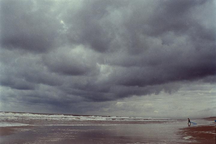 A person walks along a wet beach carrying a surfboard under a dramatic, cloudy sky.