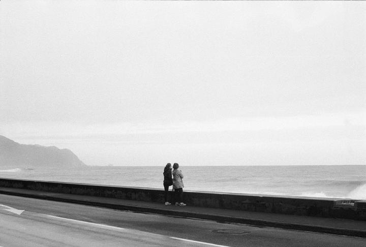 Two people stand on a promenade next to a road, looking out at the ocean with mountains in the distance.