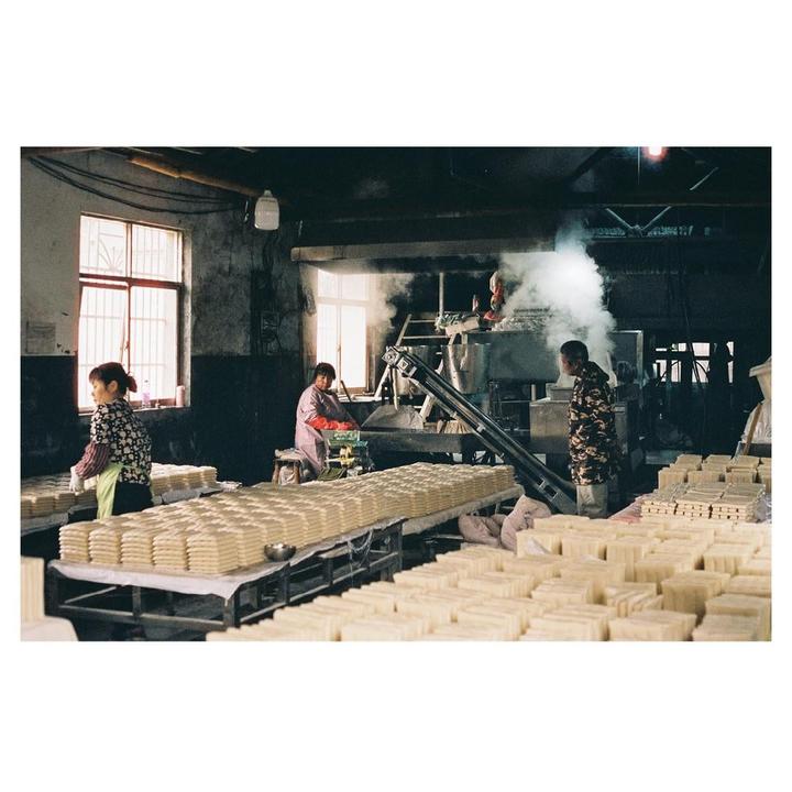 Three people work in a rice cake factory with large windows and machinery producing steam.