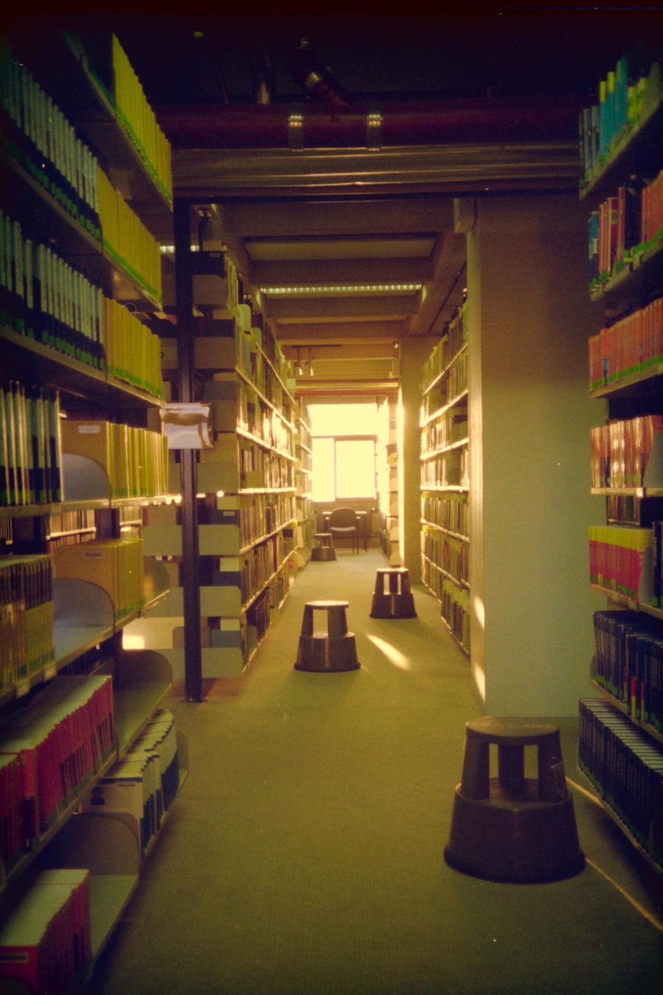 A long aisle in a library is lined with bookshelves and has three step stools on the carpeted floor, leading to a bright window at the end.