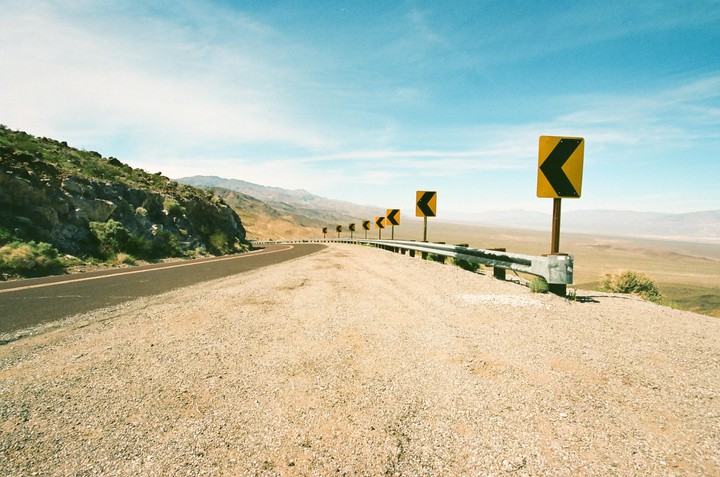 A winding desert road with a guardrail and several yellow chevron signs indicating a curve to the left, with mountains in the distance under a clear sky.