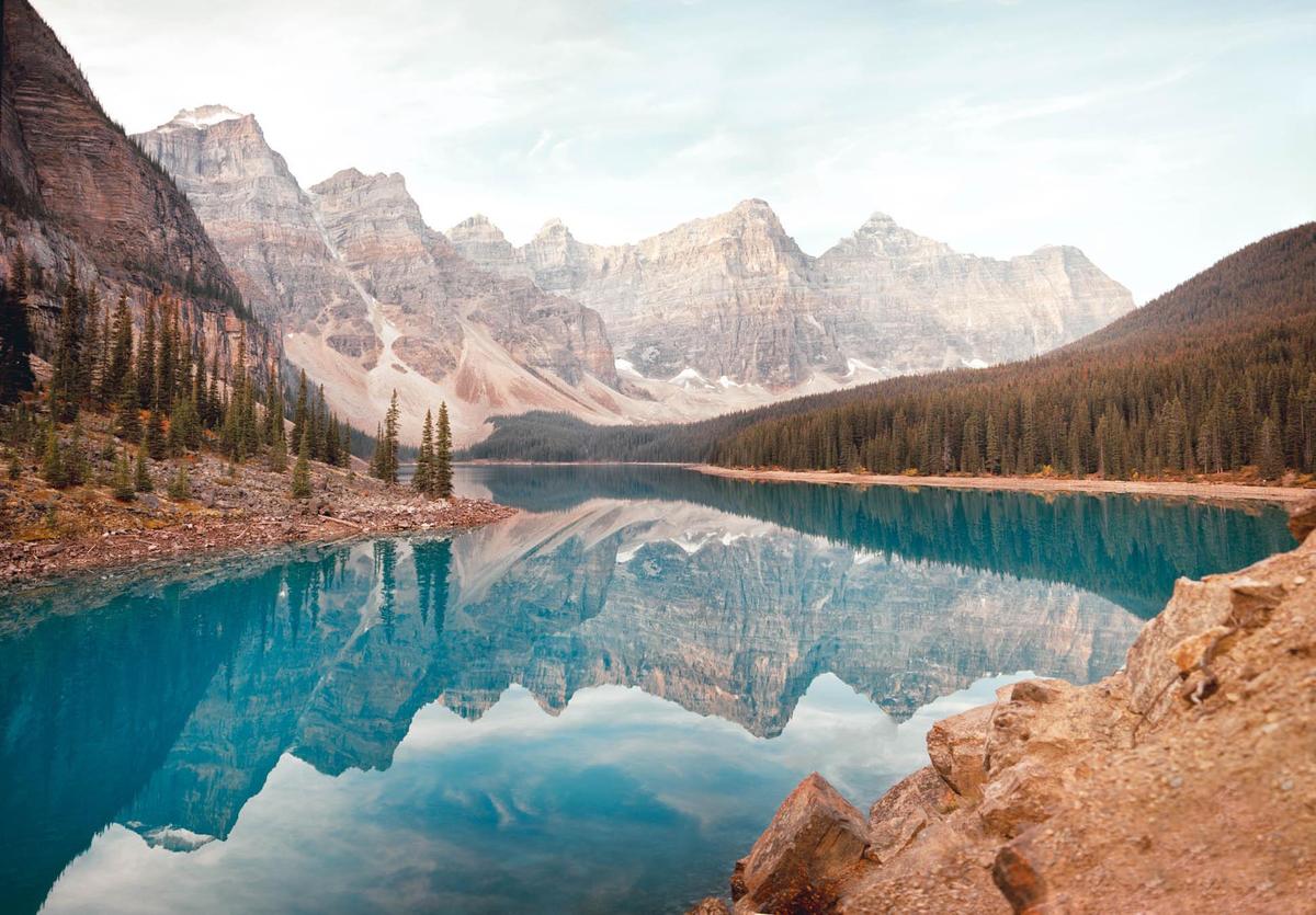 A panoramic landscape shows a turquoise lake reflecting mountains and a forest under a hazy sky.