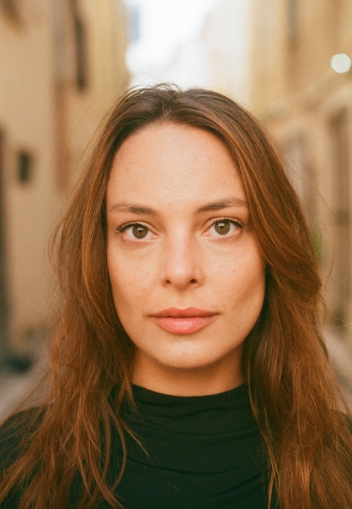 A woman with long brown hair looks directly at the viewer in a close-up portrait.