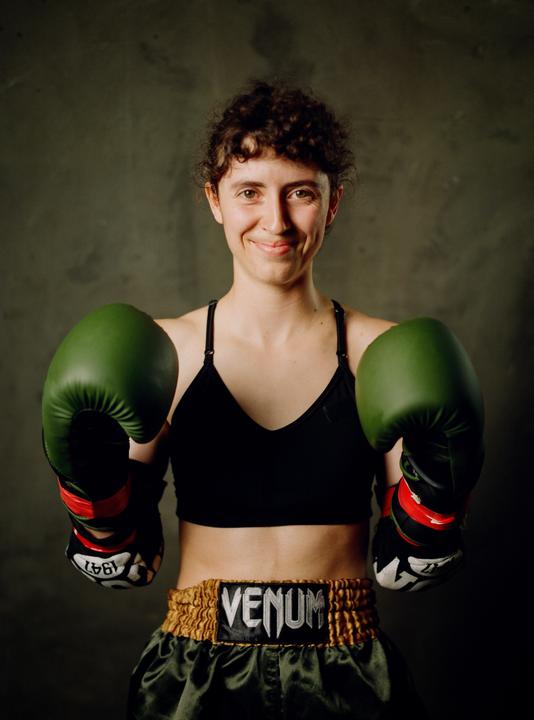 A woman wearing boxing gloves and shorts smiles at the camera.
