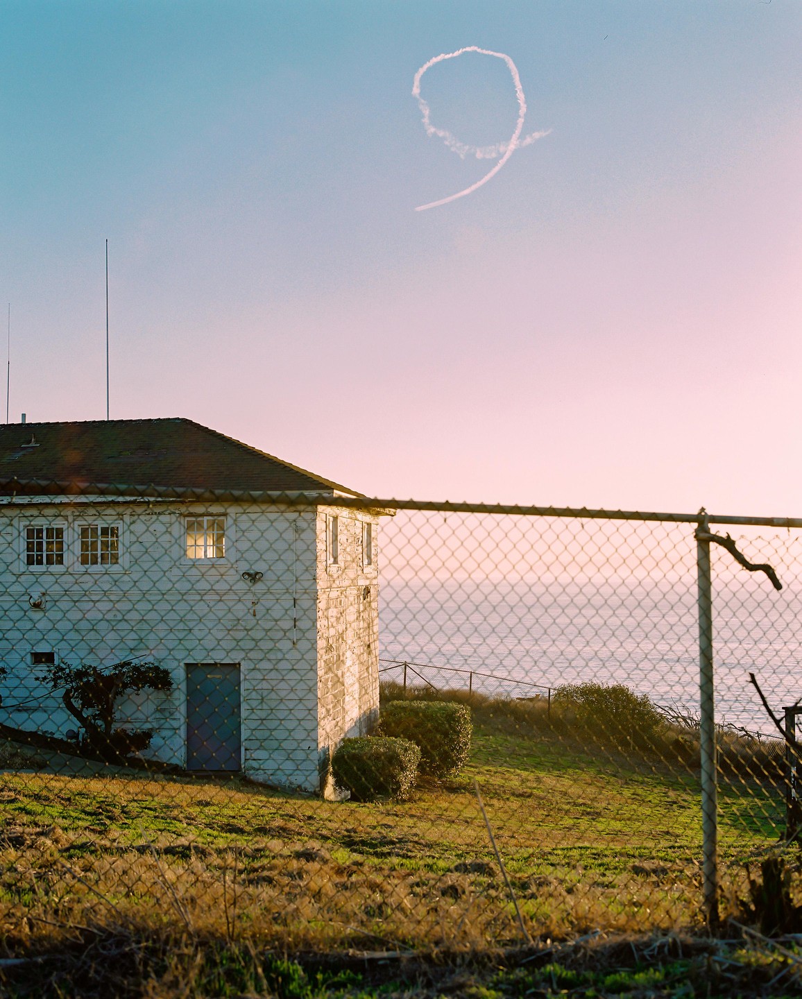 A white building with a dark roof stands on a grassy hill overlooking the ocean, with a chain-link fence in the foreground and a skywriting number nine in the sky.